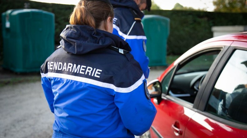 Une gendarme de dos portant un blouson de gendarmerie, en train de contrôler le conducteur d'un véhicule rouge.