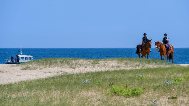 Deux gendarmes à cheval à droite sur une dune, et à gauche en mer, une vedette de la gendarmerie nationale.