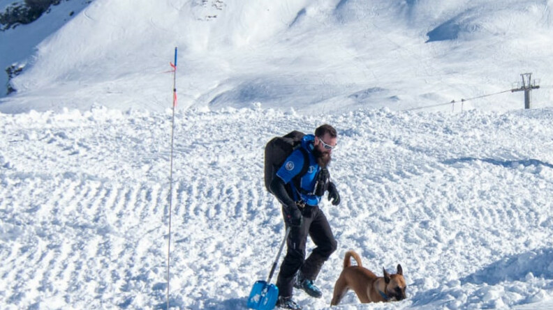 Sur une montagne enneigée, on voit une jeune homme vêtu d'une tenue de ski, équipé d'une pelle bleue, d'un gros sac à dos noir et de lunettes de ski blanches, précédé par un chien dont le museau est dirigé vers le sol.