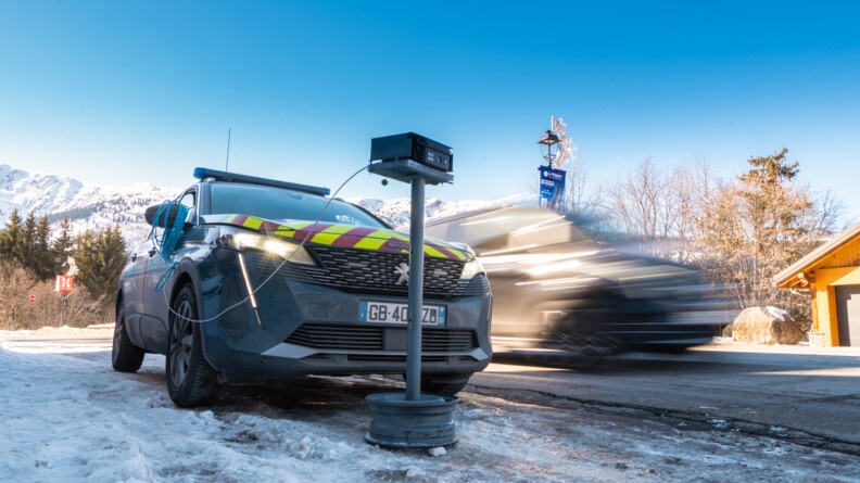 Sur la route entre Courchevel et Méribel, une voiture de gendarmerie arreté dans la neige sur le bas côté, a devant elle un drôle de radar, relié à la voiture et qui permets de vérifier les plaques des automobilistes que l'on voit passer en fond. Le ciel est bleu et les montagnes se détache en fond.