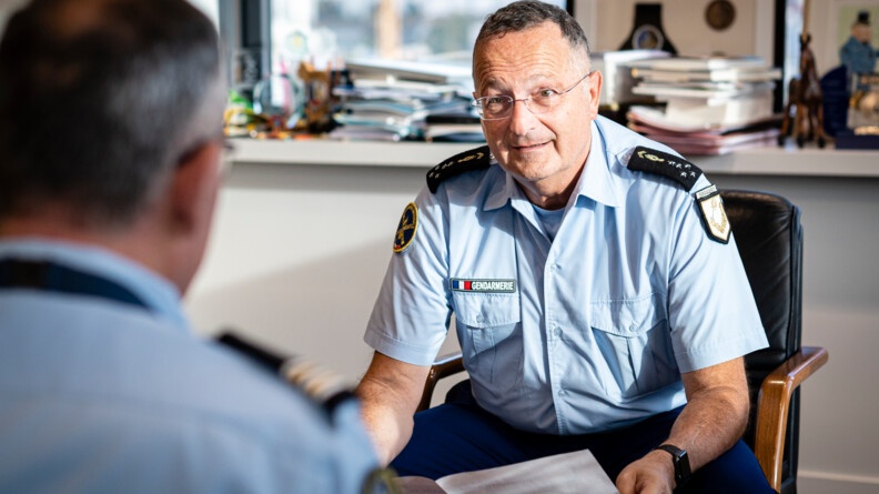 Portrait du directeur général de la gendarmerie, assis sur un fauteuil, tenant un document entre les mains. Au premier plan, face à lui, un lieutenant-colonel vu de dos.