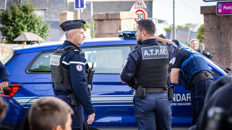 Devant un véhicule de gendarmerie, à gauche, de profil droit, un gendarme, et à droit un policier municpal en train d'échanger.