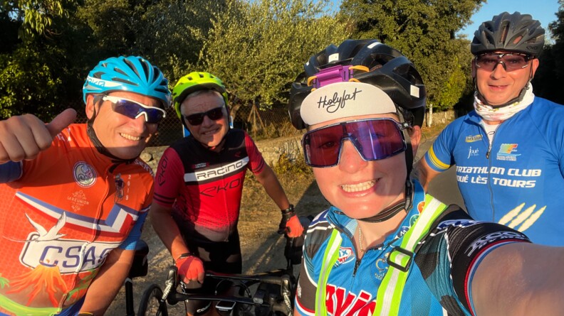 Une jeune femme cycliste, souriante, avec un casque et des lunettes, réalise un selfie avec sa main gauche. Elle est entourée de trois cyclistes hommes. L'un deux, tout à gauche de l'image, fresse son pouce.