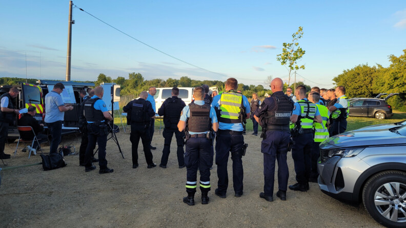 Environ 25 gendarmes regroupés en extérieur devant des véhicules et un espace boisé pour un briefing