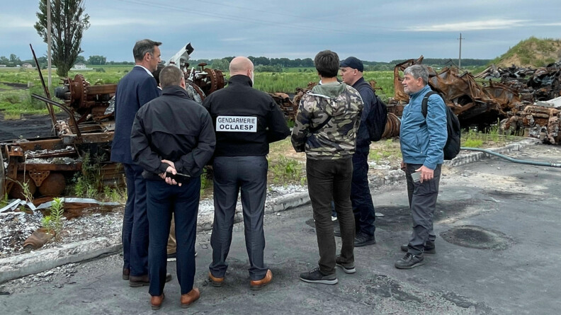 Un groupe de six hommes en civil, vus de dos ou de trois quart dos, discutent devant des carcasses de véhicules rouillées et abandonnées dans un champ.