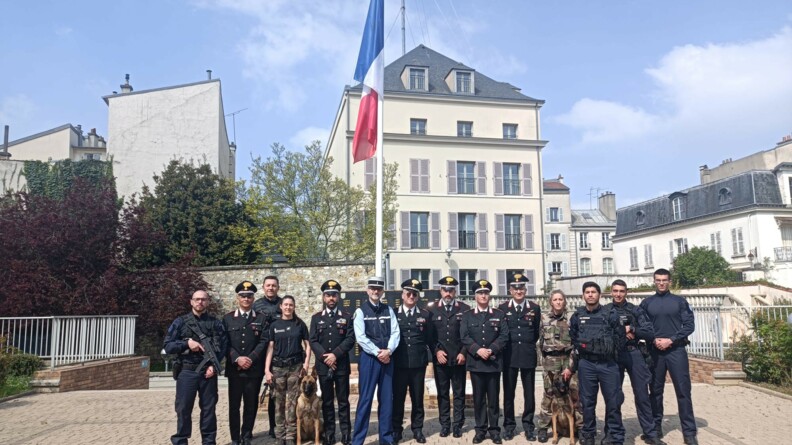 Un colonel de gendarmerie entouré de six carabiniers en tenue, de cinq militaires de PSIG et de deux maîtres de chien féminins avec leurs chiens, en rangée, sous un drapeau.