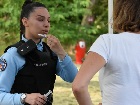 Une gendarme de face fait une démonstration de l'utilisation d'un éthylotest à une personne civile en tee-shirt blanc, vue de dos.