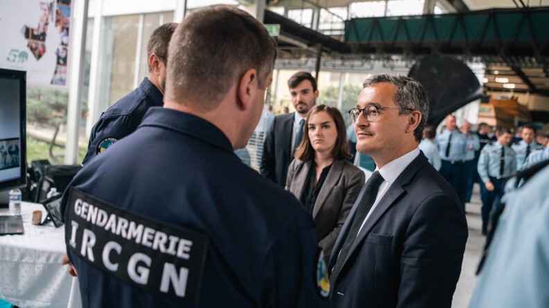 Le ministre de l'Intérieur, vu de trois-quart face, échangeant avec deux experts de l'IRCGN, dont un vu de dos au premier plan, sur le village gendarmerie.