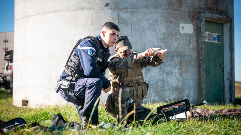 Un gendarme et un militaire de l'armée de Terre accroucpis dans l'herbe regardant dans la même direction. Le  militaire de l'armée de Terre désigne un point avec les doigts de ses deux mains.