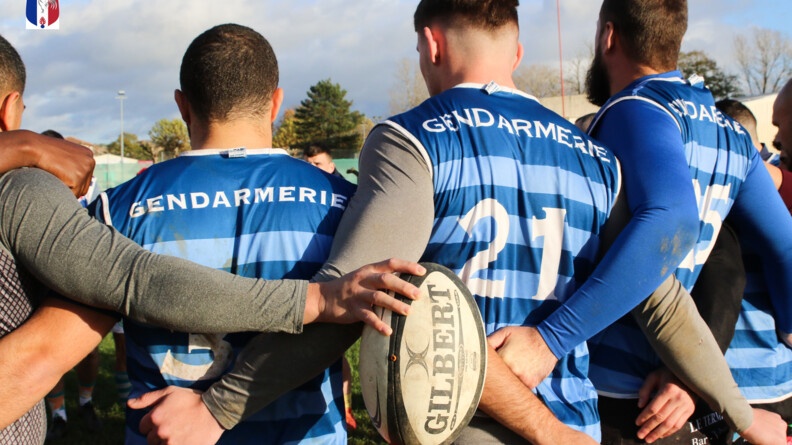 Zoom sur trois joueurs de rugby se tenant bras dessous par le bas du maillot, de dos. Au milieu de l'image, l'un d'eux tient le ballon de sa main droite