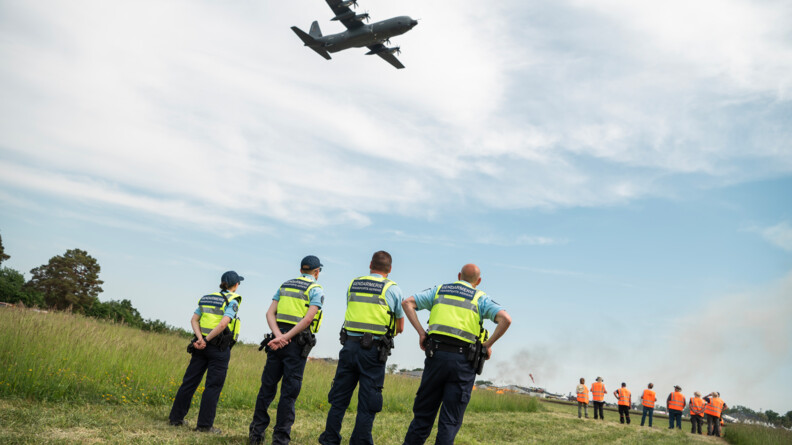 Quatre gendarmes de la GTA devant la piste de l'aérodrome. Devant eux, des personnels chargés de provoquer les explosions et au dessus d'eux, un avion passe.