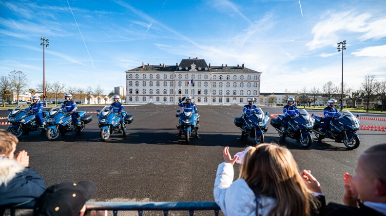 Sept motocyclistes de la garde républicaine font une démonstration devant l'école d'officiers de gendarmerie. On voit des enfants de dos applaudir au premier rang.