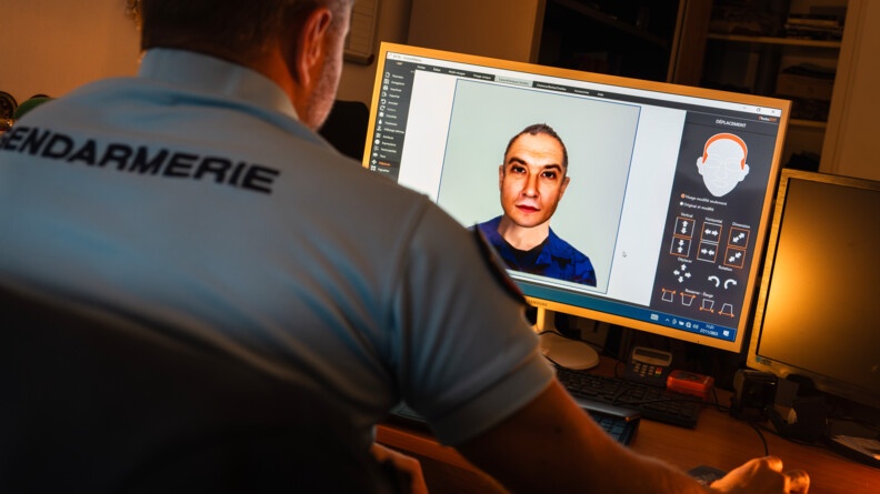 Dans une pièce sombre, on voit au pemier plan un gendarme de dos, vêtu d'un polo bleu avec l'inscription "Gendarmerie", assis devant un bureau. Face à lui, l'écran d'un ordinateur affichant le visage d'un homme d'âge moyen sur un logiciel.