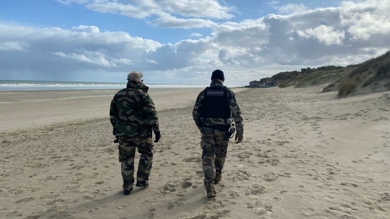 Sur la plage du nord, deux gendarmes en tenue de treillis, marchent sur la plage au bord de l'eau. Ils surveillent le littoral et l'horizon à la recherche de potentielles embarcations.