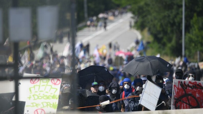 Un groupe de manifestants radicaux, masqués et casqués, portant des pancartes et des parapluies noirs.