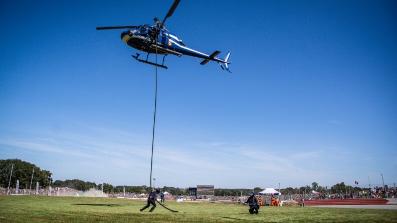 Un hélicoptère de la gendarmerie et deux militaires glissant le long de la corde sur de l'herbe. AU fond, le public