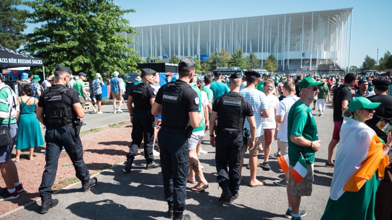 Quatre gendarmes mobiles, vus de dos, traversent une foule de supporters sur le parvis du stade de Bordeaux, que l'on voit en arrière-plan.