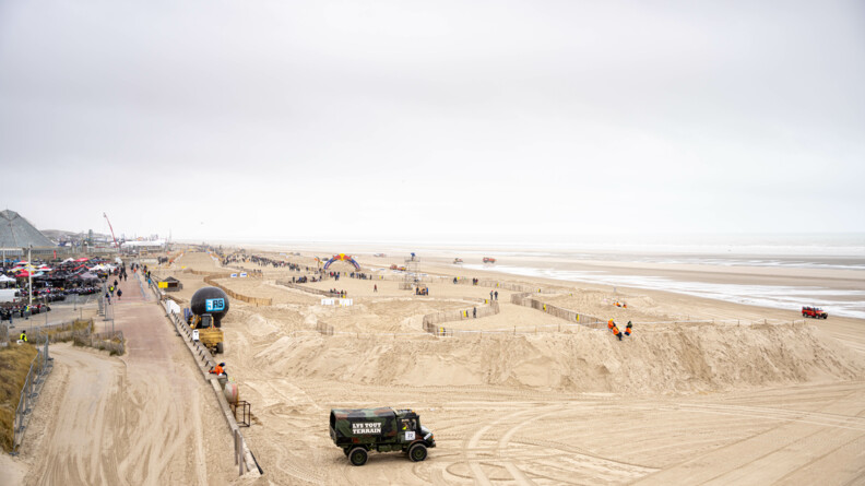 Photo du circuit de course de sable depuis les dunes. On y voit des gens, des camions. Sur la gauche on voit aussi des quads garés en attendant le début de la course.