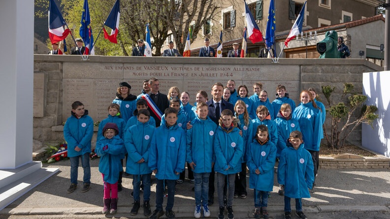 Le président de la république, Emmanuel Macron, apparait de face, debout, entouré d'enfants vêtus du même blouson bleu, devant un monument aux morts, sur lequel est inscrit "Aux enfants de Vassieux victimes de l'agression allemande morts pour la France en juillet 1944