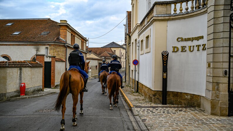Dans les rues d'un village champenois, trois gendarmes montés de la Garde républicaine avancent dans une rue, vus de dos. A droite, une entrée de maison avec l'inscription "Champagne DEUTZ3
