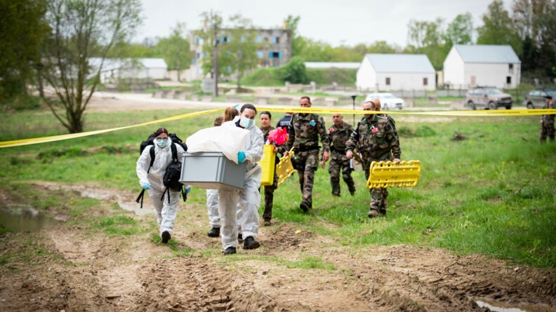 Quatre scientifiques de l'IRCGN en combinaison blanche portant du matériel dans des sacs noirs et caisses jaunes traversent sous la bande jaune gendarmerie de gel des lieux; 5 prévôts en treillis les aident à porter le matériel