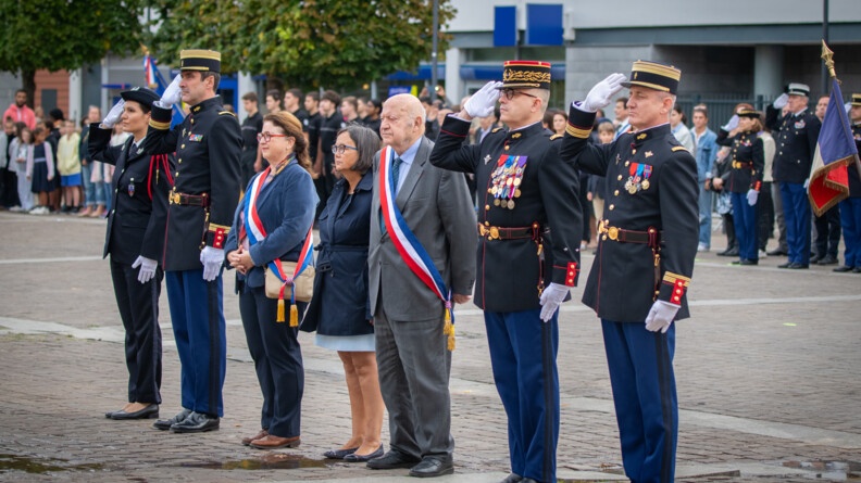 Six personnes sont alignées debout sur un parvis : quatre militaires de la garde au garde à vous, ainsi que trois civils dont deux portent en écharpe le drapeau français