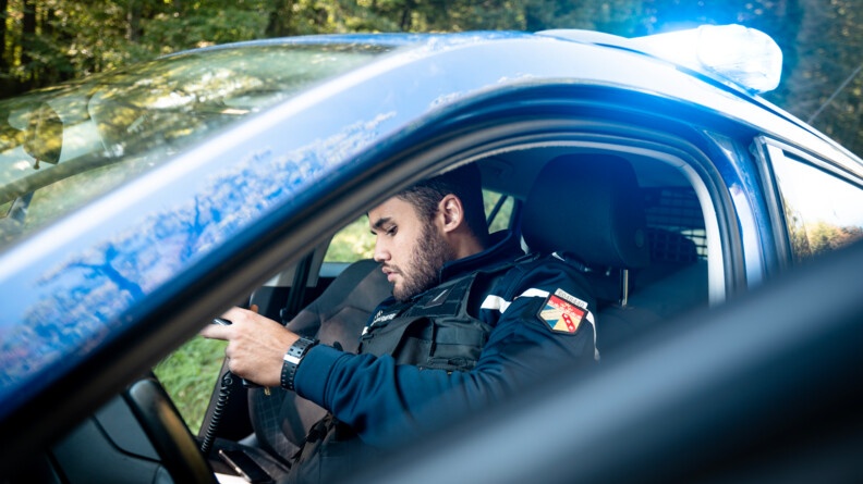 Un gendarme dans son véhicule de service, girophare allumé, assis derrière le volant, manipule sa radio.