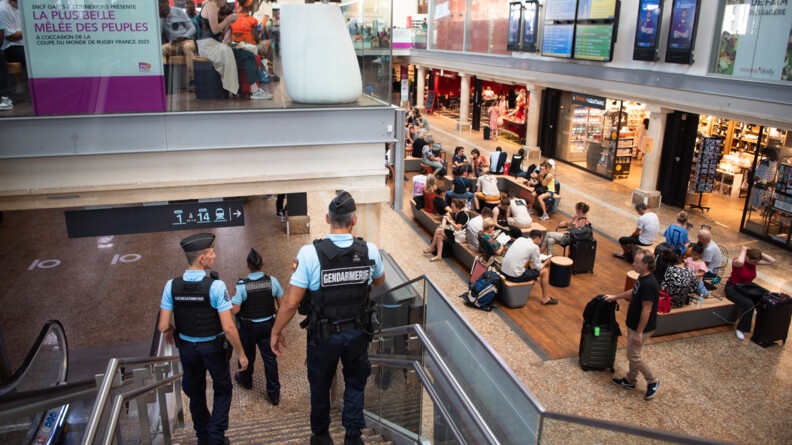 Trois gendarmes, vus de dos, en patrouille dans la gare de Bordeaux Saint-Jean, descendent des escaliers vers la salle d'attente de la gare, à droite de la photo, bordée de boutiques, avec des passagers qui attendent. En haut à gauche, la salle d'attente de l'étage.