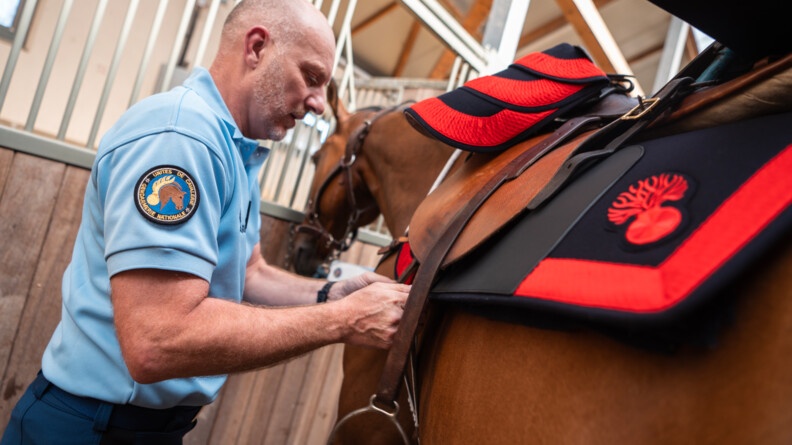 Un gendarme avec l'écusson de la Garde républicaine, en train d'ajuster la selle au tapis noir et rouge de son cheval