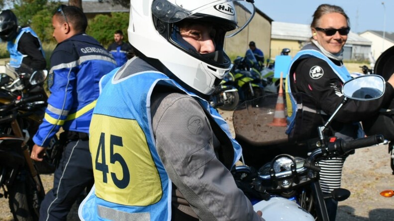 Motocycliste civil casqué, portant un dossard bleu et jaune numéroté 45, à cheval sur sa moto. En arrière-plan, à droite, une autre motocycliste, non casquée, et à gauche, un motocycliste de l'EDSR 78 de trois-quart dos.
