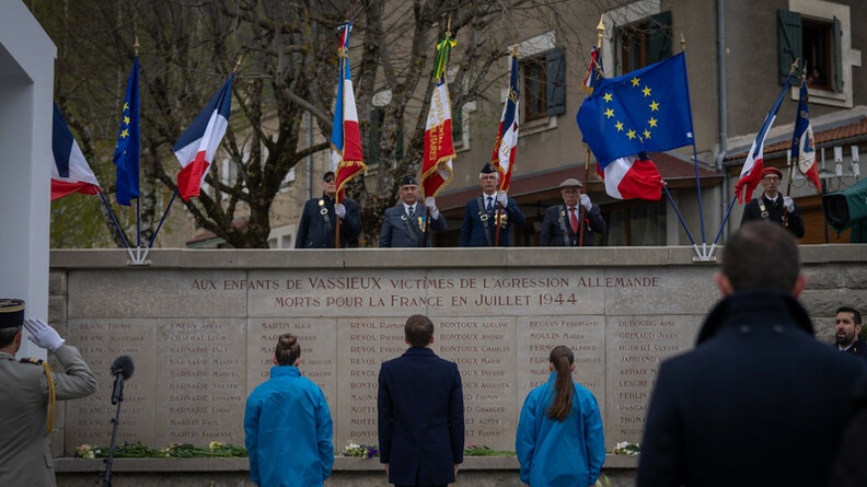 Le président de la république, Emmanuel Macron, apparait de dos, debout, entouré de deux jeunes filles vêtues du même blouson bleu, devant un monument aux morts, sur lequel est inscrit "Aux enfants de Vassieux victimes de l'agression allemande morts pour la France en juillet 1944