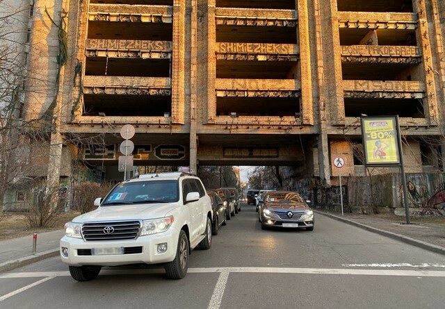 Un véhicule blindé du GIGN de couleur blanche ouvre la voie à un convoi de véhicules sombres sur deux colonnes passant sous un bâtiment.