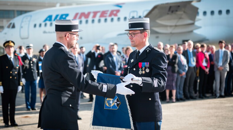 Au premier plan,  le colonel Philippe Mirabaud, à droite, reçoit le fanion de la GTA des mains de son précédesseur, lors de sa prise de commandement, à l'été 2022. Au second plan, un parterre de personnes, civiles et militaires, et un avion sur le tarmac.