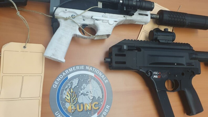 Deux armes fabriquées via une imprimante 3D et posées sur une table en bois avec l'écusson de l'unité nationale cyber.