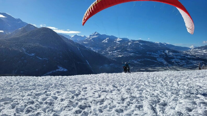 Un parapente décolle sur un sol enneigé, dans un paysage montagneux. Le ciel est bleu et le paysage est ensoleillé