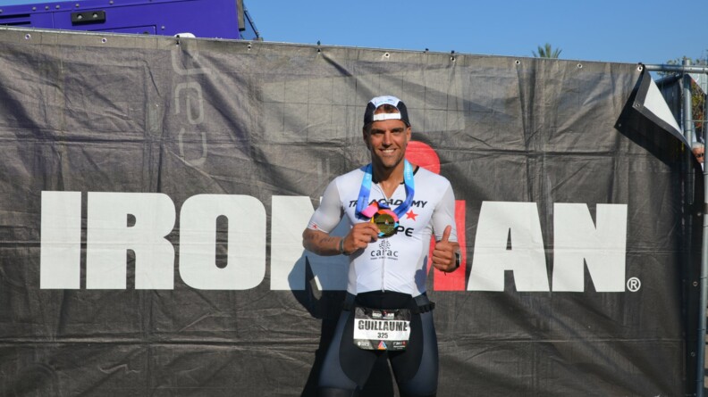 Le MDC Guillaume, revêtu d'un cycliste noir, d'un maillot blanc et casquette sur la tête, pose devant un grand panneau noir portant l'inscription IRONMAN en blanc et montre, en souriant, sa médaille de bronze.