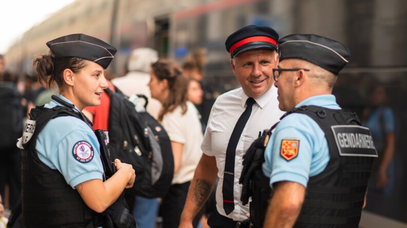 Une réserviste et un réserviste de la gendarmerie, vus de trois-quart, en discussion avec un contrôleur, vu de face, sur le quai de la gare.