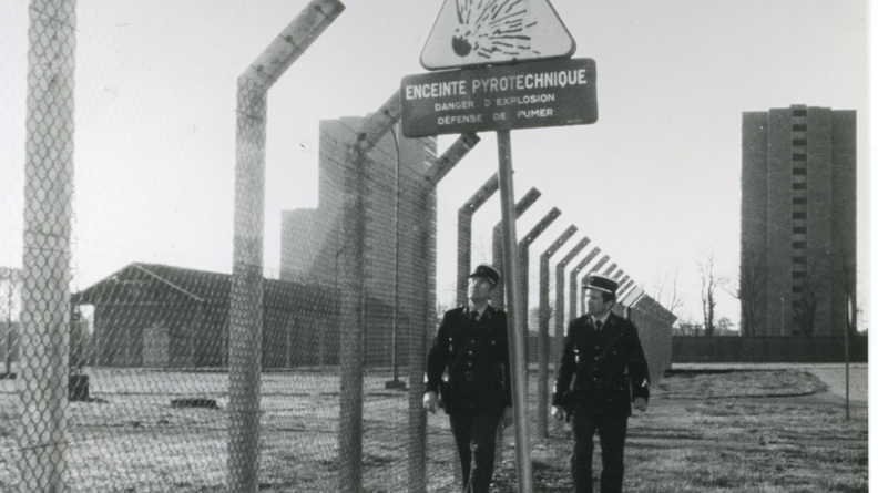 Sur une photo en noir et blanc, deux gendarmes marchent le long d'un grillage représentant de toute évidence la bordure d'un site militaire dont ils ont en charge la sécurisation.