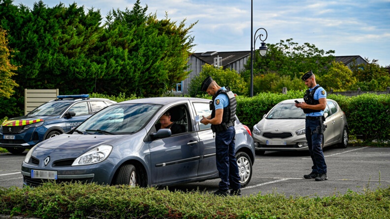 Sur un petit parking, deux gendarmes mobiles, sur la partie droite de l'image, procédent au contrôle d'une conductrice d'un véhicule léger gris.