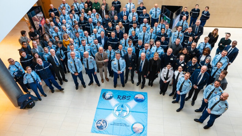 Dans un vaste espace intérieur, de nombreux gendarmes en tenue formant un demi-cercle sont photographiés par en haut. Tous lèvent la tête en direction de l'objectif. Au sol est disposé un grand tissu rectangulaire carré de couleur bleu ciel sur lequel il est inscrit en gros, en bleu marine RBS.
