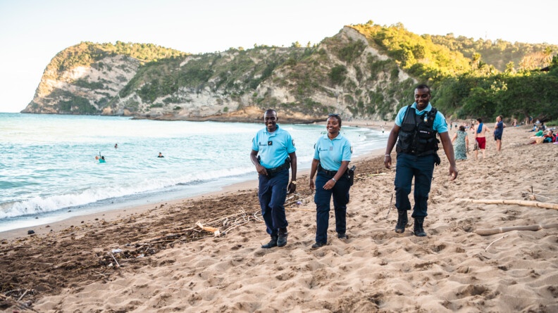 Trois gendarmes de face marchent le long d'une plage où se trouvent des baigneurs.