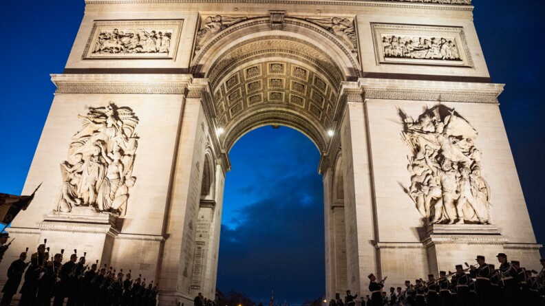 Vue en contre-plongée sur l'Arc de Triomphe éclairé, tandis que la nuit est tombée et l'on distingue des troupes rassemblées de part et d'autre et des personnes sous le monument.