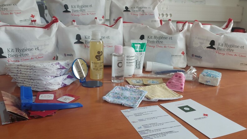 Sur une table 8 kits "hygiène et bien-être" avec le type de contenu: peigne, miroir, gel douche, crème hydratante, cotons démaquillants, mouchoirs, serviettes hygiéniques