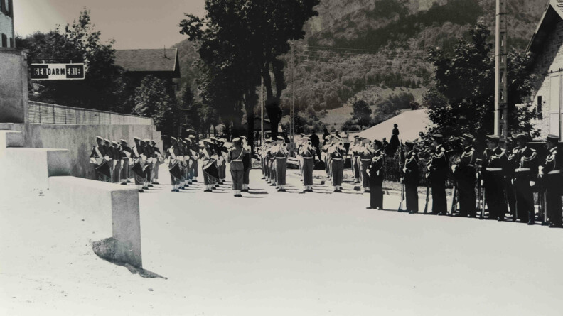 La photo, en noir et blanc, représente des militaires debout, alignés, en plusieurs rangées, face à un bâtiement de gendarmerie, quie l'on devine, et dont une voit la pancarte. On aperçoit en arrière-plan le flan d'une colline ainsi qu'une paroi rocheuse