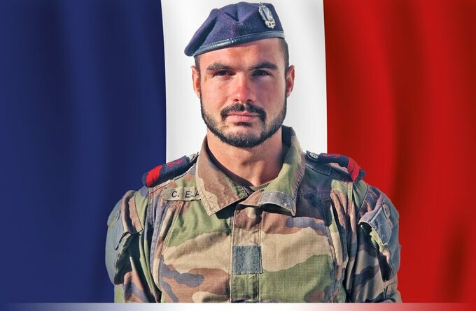Un jeune militaire brun, yeux marrons, portant une barbe, pose en tenue kakie devant un drapeau bleu blanc rouge recouvrant intégralement le fond de l'image.