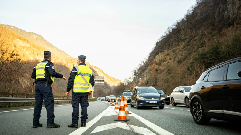 Deux gendarmes avec une chasuble jaune  vérifient l'état du trafic sur la route. A droite, on voit une colonne de voitures. Au fond, les montagnes.