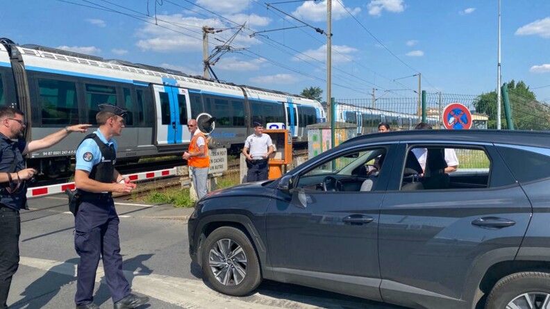 Un gendarme dispense des conseils de prévention à un conducteur arrêté à un passage à niveau, tandis qu'un train passe. Autour, d'autres agents de l'Etat et de la SNCF sont présents.