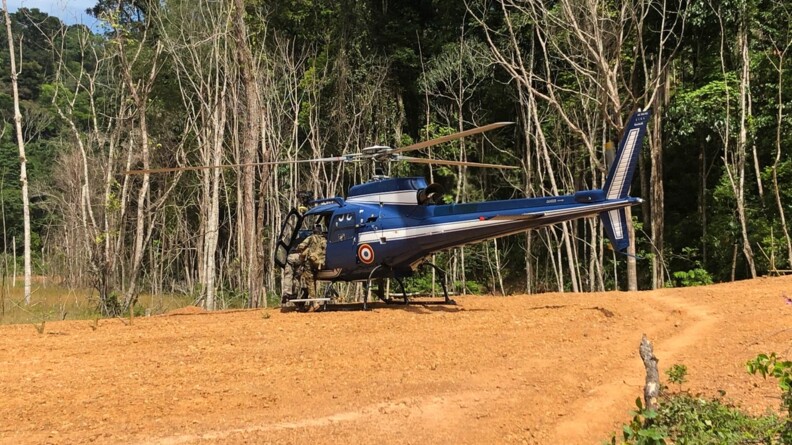 Un militaire de la gendarmerie en treillis kaki montant à bord d'un hélicoptère bleu de la section aérienne de gendarmerie de Cayenne posé en lisière de forêt, sur un terrain ocre.