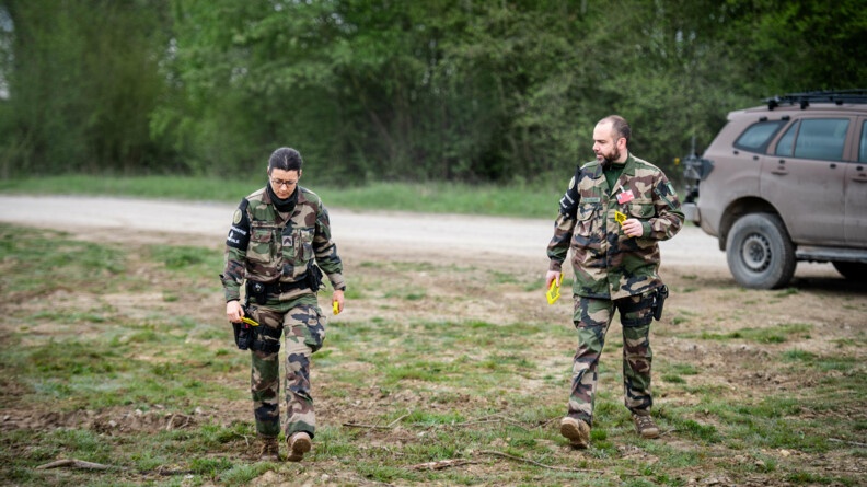 Deux gendarmes en treillis sont sur le bord d'une route en terre dans la forêt. Ils tiennent des flèches jaunes dans les mains, elles servent à montrer le chemin vers le charnier.