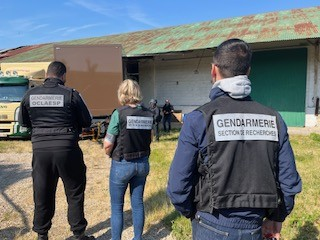 Trois enquêteurs de la gendarmerie, en civil, portant pour deux d'entre eux la chasuble de la section de recherches et pour le troisième celle de l'OCLAESP. Ils sont vus de dos, à l'extérieur d'un grand hangar, devant lequel est stationné un poid-lourd.